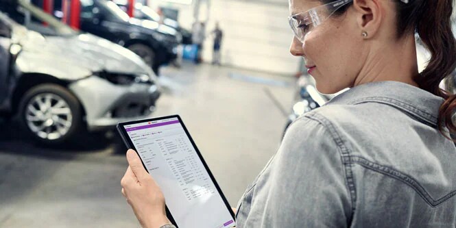 A woman standing in an autobody shop looks at the screen of a tablet device.
