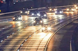 A four-lane highway at night has many cars with bright headlights.