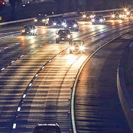 A four-lane highway at night has many cars with bright headlights