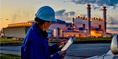 man in a construction hat on an ipad in front of an industrial plant
