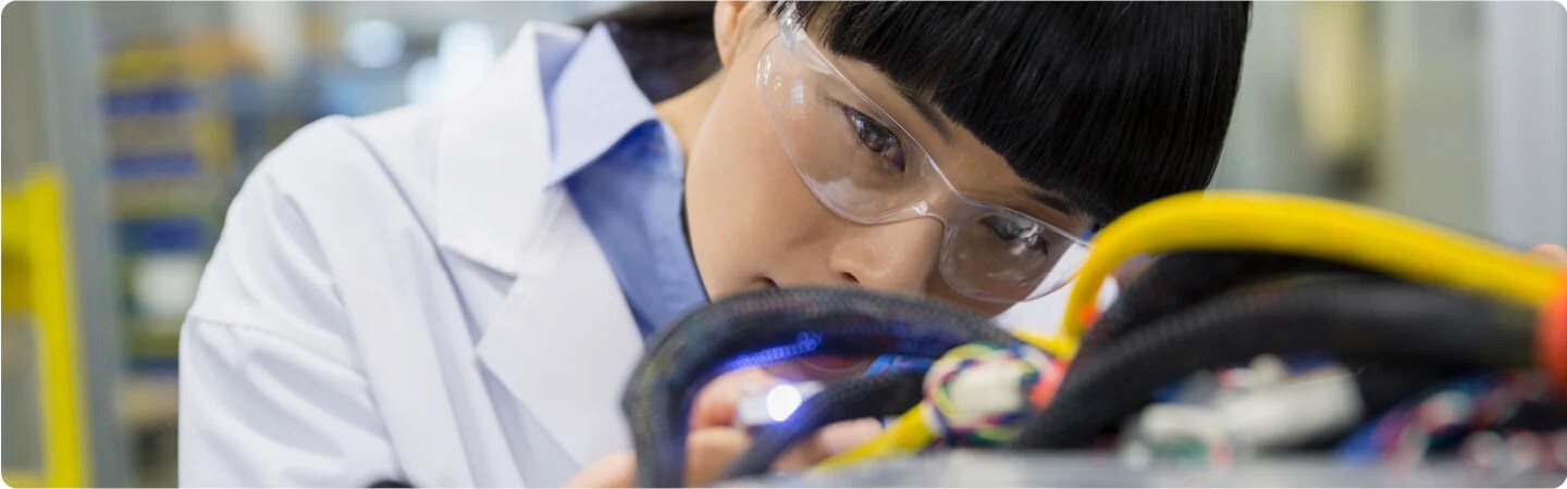 A close up of a person wearing safety glasses and a lab coat looks closely at tubing and electrical wires.

