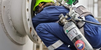 Person in full protective suit and helmet descending into a large industrial pipe using a safety harness and rope during a confined‑space operation.
