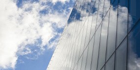 Exterior photo looking up at glass wall building against blue sky with clouds
