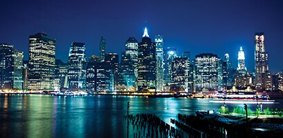 A city landscape with a body of water in the forefront, taken at night with lights illuminating the buildings and reflections of the city on the water.
